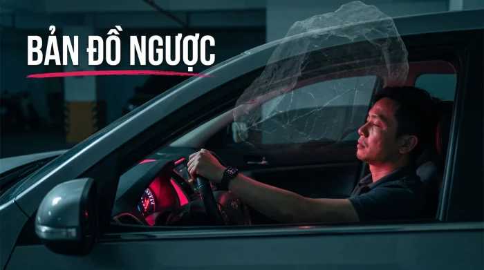 Người đàn ông ngồi ghế lái trong gara tối, mắt nhắm, kiệt sức; một tảng đá bán trong suốt đè trên nóc xe, bảng điều khiển hắt ánh hồng đỏ lên mặt, kèm chữ “BẢN ĐỒ NGƯỢC”.